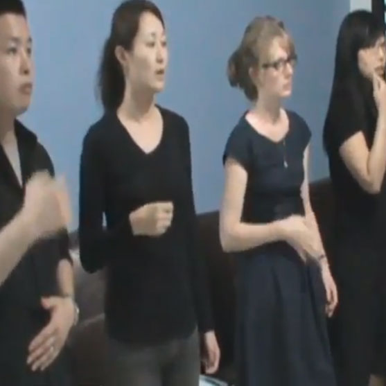 Four people in dark clothing standing side by side against a blue wall, with hands raised in a gesture.