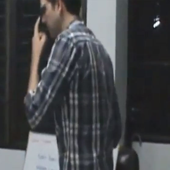 A man in a plaid shirt stands indoors near a window with his hand on his face.