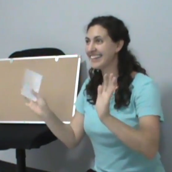 Smiling woman in a light blue shirt gesturing with her hands while standing beside a bulletin board.