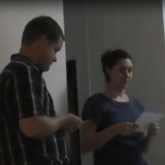 A man and a woman stand indoors and look at papers while discussing them.