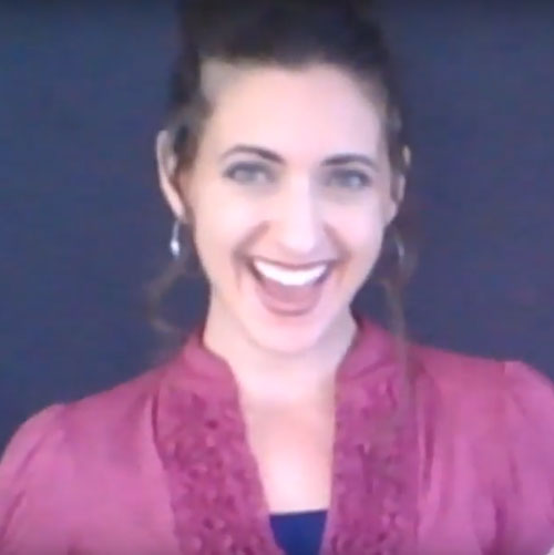A smiling woman wearing a pink blouse and earrings stands against a dark background.