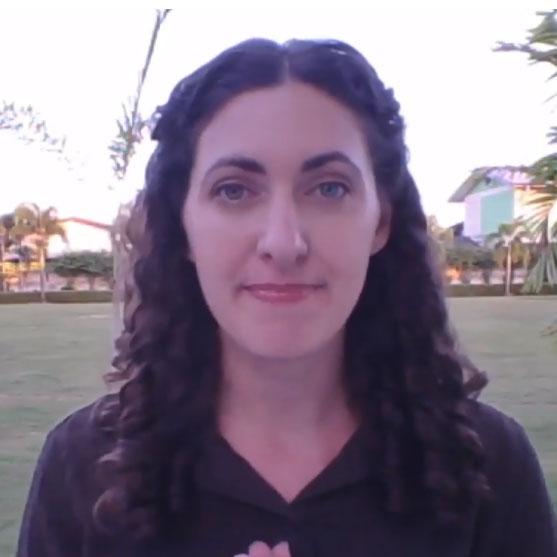 Woman with dark curly hair standing in a grassy outdoor area with trees and buildings in the background.