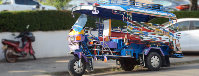 A brightly decorated three-wheeled tuk-tuk parked or driving along a street, with cars and a motorcycle in the background.