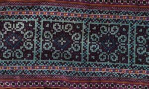 A colorful woven textile with repeating geometric borders and ornate square motifs in dark purple, teal, red, and white.